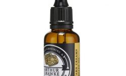 A short cylindrical brown glass bottle with a black rubber pipette lid and a black label that has Arthur J Hawke Custard Creams Beard Oil written in bold white writing on it, on a white background.