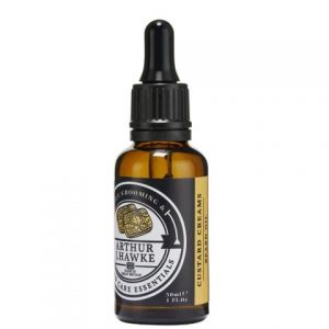 A short cylindrical brown glass bottle with a black rubber pipette lid and a black label that has Arthur J Hawke Custard Creams Beard Oil written in bold white writing on it, on a white background.