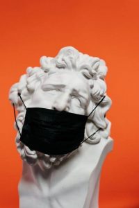 A large marble bust of a persons head wearing a black medical mask, on an orange background.