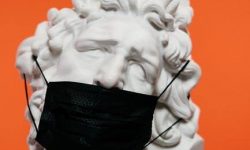A large marble bust of a persons head wearing a black medical mask, on an orange background.