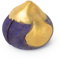 A large dark purple fig shaped bubble bar thats covered in some gold glitter and has a cut in the middle of it that's filled with some bright white cocoa butter, on a white background.