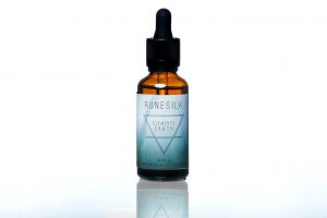A short cylindrical brown glass bottle with a black rubber pipette lid and a white and gradient green label that has runesilk elements earth beard oil written in bold black writing and a picture of a black triangle on it, on a white background.