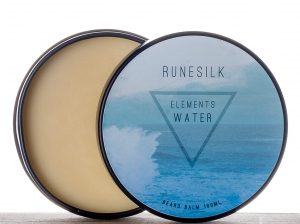 A short cylindrical silver open tin full of some golden yellow beard balm with a white and gradient blue label that has Runesilk Elements Water Beard Balm written in bold black writing and a picture of a blue triangle on it, on a white background.