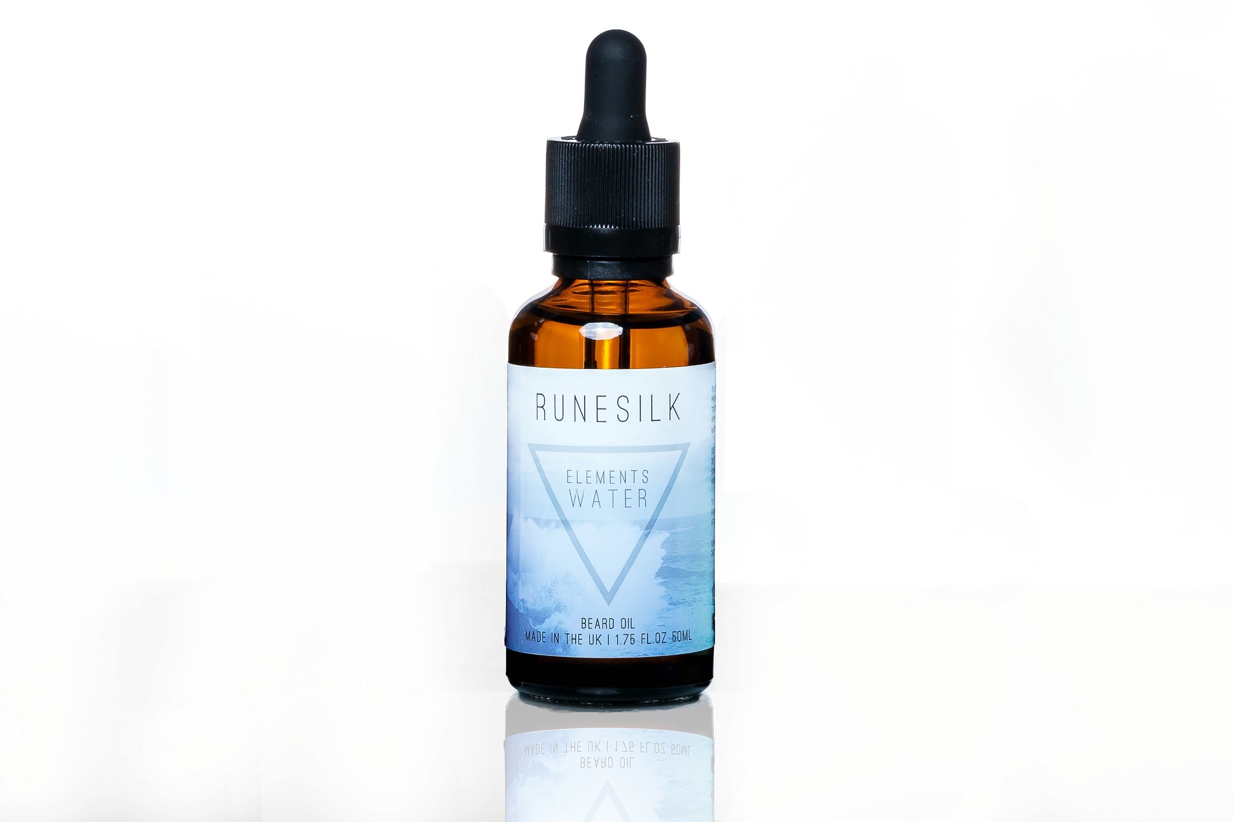 Finding Water At The Bottom Of My Beard | Runesilk Elements Water Beard Balm & Beard Oil A short cylindrical brown glass bottle with a black rubber pipette lid and a white and gradient blue label that has runesilk elements earth beard oil written in bold black writing and a picture of a blue triangle on it, on a white background.