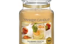 A tall glass jar full of some creamy yellow wax with a label that has Yankee Candle written in black writing, Calamansi Cocktail written in black writing, and a picture of a cocktail glass that has a slice of orange and a slice of lime inside it and a sprig of Mint coming out of the top of it on it, on a white background.