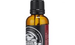 A short cylindrical dark green glass bottle with a black rubber pipette lid and a black label that has Arthur J Hawke Cherry Cola Beard Oil written in bold white writing on it, on a white background.