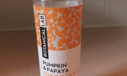 A tall cylindrical clear plastic bottle full of some clear liquid with a bright orange a white gradient label that has Botanical Lab Daily Exfoliator written in bold black writing and Pumpkin & Papaya Enzyme written in bold orange writing on it, on a white background.