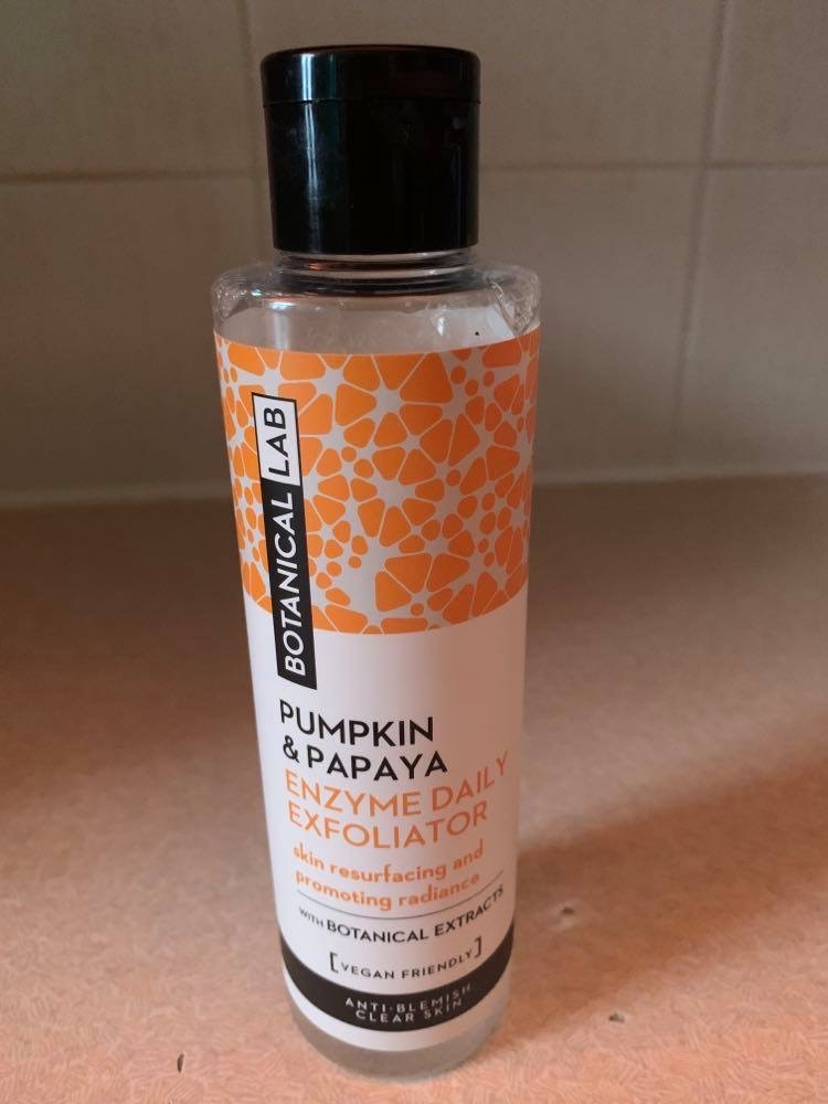 A tall cylindrical clear plastic bottle full of some clear liquid with a bright orange a white gradient label that has Botanical Lab Daily Exfoliator written in bold black writing and Pumpkin & Papaya Enzyme written in bold orange writing on it, on a white background.