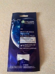 A long thin rectangular dark blue plastic pouch that has Garnier Moisture Bomb Night Time Eye Tissue Mask written in bold white writing and a picture of a droplet of Water on it, on a white background.