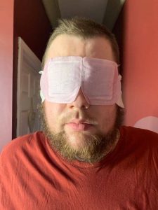 A tall bearded man wearing a dark red T-shirt and a bright pink eye mask that has some white paper arms attached to it stood against a dark burgundy wall.