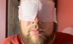 A tall bearded man wearing a dark red T-shirt and a bright pink eye mask that has some white paper arms attached to it stood against a dark burgundy wall.