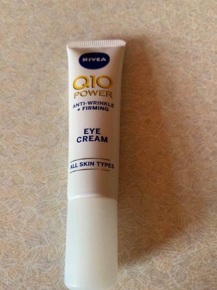 The Power Of A Great Eye Cream | Nivea Q10 Power Anti-Wrinkle + Firming Eye Cream A short flat conical white plastic tube that has Nivea written in bold blue writing and Q10 Power Anti-Wrinkle + Firming Eye Cream written in bold yellow writing on it, on a white background.