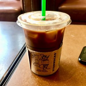 A tall cylindrical clear plastic cup with a clear plastic lid full of some dark brown and cream liquid that has a brown cardboard sleeve around it and a green plastic straw coming out of the top of it on a rectangular light wooden table, on a white background.
