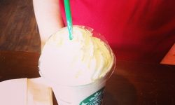A tall cylindrical clear plastic cup with a clear plastic lid full of some bright white strawberry milkshake, some pieces of strawberries and some strawberry syrup with a green plastic straw coming out of the top of it on a light wooden table, on a white background.