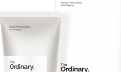 A short flat conical white plastic tube with a white plastic lid and a white label that has The Ordinary Natural Moisturising Factors + HA written in bold black writing next to a tall rectangular cardboard box that has The Ordinary Natural Moisturising Factors + HA written in bold black writing on it, on a white background.