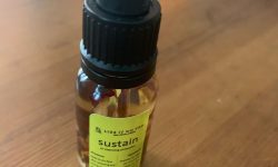 A short and thin cylindrical clear glass bottle full of some clear liquid and pink rose petals with a black rubber pipette lid and a white label that has Like It On Top Sustain Oil Cleansing Protection written in bold black writing on it, on a white background.