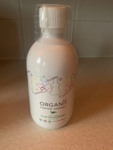A tall wide cylindrical clear plastic bottle full of some clear liquid with a clear plastic measuring cup lid that has Organii written in bold black writing and Aloe Vera Eucalyptus Mouthwash written in bold Green writing on it, on a white background.