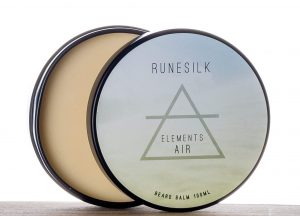  A short cylindrical silver open tin full of some golden yellow beard balm with a white and gradient light blue label that has Runesilk Elements Air Beard Balm written in bold black writing and a picture of a light blue triangle on it, on a white background.