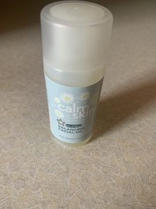A short cylindrical light green plastic Bottle that has Superdrug written in bold black writing, Calm Skin De-Stress Balancing Facial Oil written in bold white writing, and some pictures of some white chamomile petals on it, on a white background.
