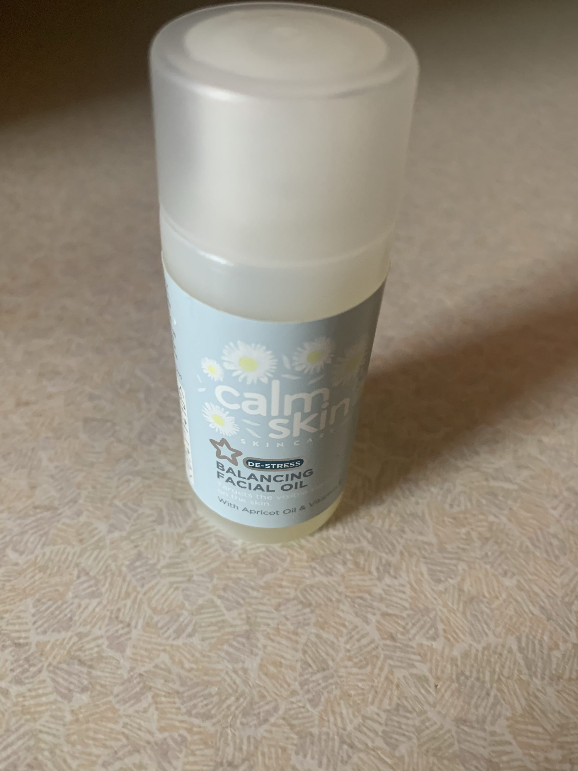 A short cylindrical light green plastic Bottle that has Superdrug written in bold black writing, Calm Skin De-Stress Balancing Facial Oil written in bold white writing, and some pictures of some white chamomile petals on it, on a white background.