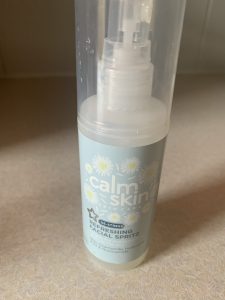 A tall cylindrical light green plastic spray bottle that has Superdrug written in bold black writing, calm skin de-stress refreshing Facial Spritz written in bold white writing, and some pictures of some white chamomile flowers on it, on a white background.