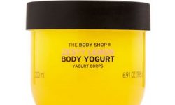 A short rounded rectangular clear plastic tub full of some bright yellow liquid with a dark green lid and a bright yellow label that has The Body Shop Zesty Lemon Body Yoghurt written in dark green writing and some pictures of some bright yellow Lemons on it, on a white background.