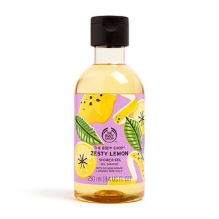 A tall cylindrical clear plastic bottle full of some bright yellow liquid with a dark green lid and a bright yellow label that has The Body Shop Zesty Lemon Shower Gel written in dark green writing and some pictures of some bright yellow lemons on it, on a white background.