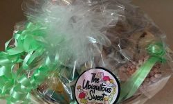 A large oval brown wicker basket full of several packets of sweets all covered in a thick layer of clear cellophane and tied up with several pieces of bright pink ribbon, on a white background.