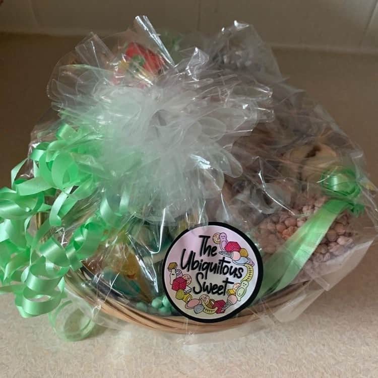 What Kind Of Candy Do You Want? | The Ubiquitous Sweet Pick 'N' Mix Sweet Hamper | Gifted A large oval brown wicker basket full of several packets of sweets all covered in a thick layer of clear cellophane and tied up with several pieces of bright pink ribbon, on a white background.