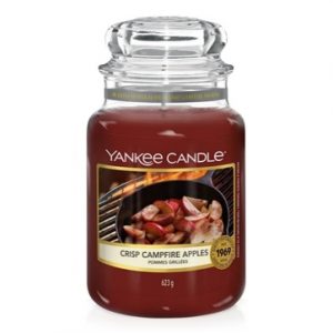 A tall glass jar full of some dark red wax with a label that has Yankee Candle written in black writing, crisp campfire apples written in black writing, and a picture of some apples being cooked in a frying pan on an open fire on it, on a white background.