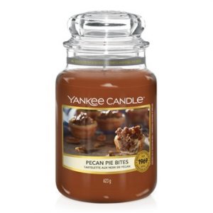 A tall glass jar full of some dark orange wax with a label that has Yankee Candle written in black writing, Pecan Pie Bites written in black writing, and a picture of a large cake that has some pecans on top of it on it, on a white background.