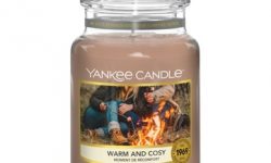 A tall glass jar full of some sandy yellow wax with a label that has Yankee Candle written in black writing, Warm & Cosy written in black writing, and a picture of some people sat around a bonfire toasting some marshmallows on it, on a white background.