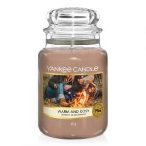 A tall glass jar full of some sandy yellow wax with a label that has Yankee Candle written in black writing, Warm & Cosy written in black writing, and a picture of some people sat around a bonfire toasting some marshmallows on it, on a white background.