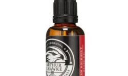 A short cylindrical brown glass bottle with a black rubber pipette lid and a white label that has Arthur J Hawke Jam Doughnut Beard Oil written in bold white writing and a picture of a black and white hawk on it, on a white background.