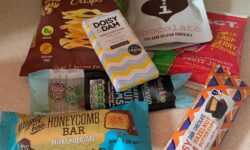 A selection of several different food packets including a large rectangular crisp packet, two small rectangular pouches, two thin rectangular pouches and a chocolate bar on top of a large rectangular piece of clear plastic, on a white background.