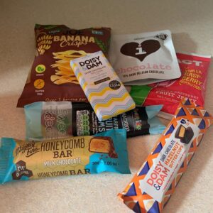 A selection of several different food packets including a large rectangular crisp packet, two small rectangular pouches, two thin rectangular pouches and a chocolate bar on top of a large rectangular piece of clear plastic, on a white background.