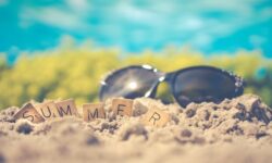 A large pair of dark black sunglasses next to a large rectangular wooden board that has summer written in bold black writing on it on a golden sandy beach.
