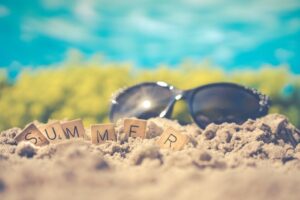 A large pair of dark black sunglasses next to a large rectangular wooden board that has summer written in bold black writing on it on a golden sandy beach.