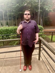 A tall bearded man wearing a pair of silver and black aviator sunglasses with some green lenses, a dark purple and white dot print shirt, a pair of Black Corduroy Trousers and a pair of shiny bright red shoes holding a bright red cane stood against a bright silver railing, on a white background.