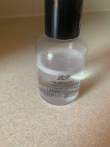 A short cylindrical clear plastic bottle full of some clear liquid with a black plastic Lid and a black label that has Boots Ingredients Glycolic Acid Toner written in bold white writing on it, on a white background.