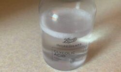 A short cylindrical clear plastic bottle full of some clear liquid with a black plastic Lid and a black label that has Boots Ingredients Glycolic Acid Toner written in bold white writing on it, on a white background.