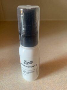 A short cylindrical black plastic bottle with a black plastic pump lid and a white label that has Boots Ingredients Hyaluronic Acid Serum written in bold black writing on it, on a white background.