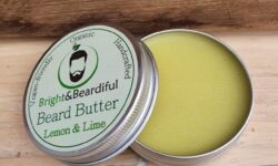 A short cylindrical open metallic Silver tin full of some creamy white beard butter with a white label that has Bright & Beardiful Lemon & Lime Beard Butter written in bold black writing and a picture of a person with a black beard on it, on a white background.
