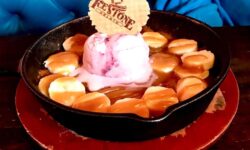 A large dark brown skillet full of some dark golden brown cookie dough, some slices of bright yellow banana, some golden brown toffee sauce, a pile of pale red gelato and a large circular golden brown wafer that has icestone engraved on it on a light wooden table, on a white background.