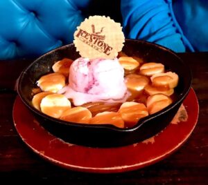 A large dark brown skillet full of some dark golden brown cookie dough, some slices of bright yellow banana, some golden brown toffee sauce, a pile of pale red gelato and a large circular golden brown wafer that has icestone engraved on it on a light wooden table, on a white background.