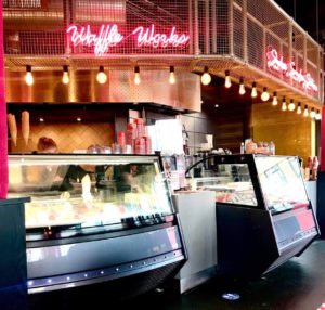 A large rectangular silver counter next to a large rectangular glass cabinet that has some small circular white bowls full of some creamy white gelato inside it and waffle wacko written in bright pink neon lights above it, on a white background.