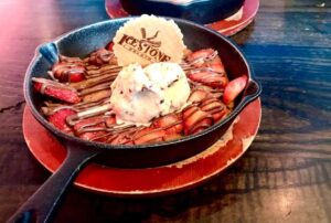 A large dark brown skillet full of some dark golden brown cookie dough, some slices of dark red strawberries, some dark brown chocolate sauce, a pile of creamy white gelato that has some pieces of crushed up kinder bueno in it and a large circular golden brown wafer that has icestone engraved on it on a light wooden table, on a white background.