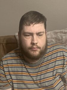 A tall bearded man wearing a dark grey and mustard yellow t-shirt sat on a dark brown leather sofa.