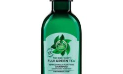 A tall cylindrical clear plastic bottle full of some dark green liquid with a black plastic lid and a dark green label that has The Body Shop Fuji Green Tea™ Refreshingly Purifying Shampoo written in bright green writing and some pictures of some bright green tea leaves on it, on a white background.