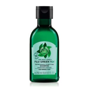 A tall cylindrical clear plastic bottle full of some dark green liquid with a black plastic lid and a dark green label that has The Body Shop Fuji Green Tea™ Refreshingly Purifying Shampoo written in bright green writing and some pictures of some bright green tea leaves on it, on a white background.